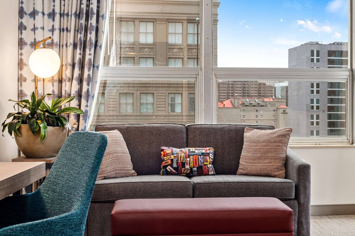 Una sala de estar moderna con un sofá gris, un cojín colorido, un sillón azul y un ventanal con vistas a la ciudad.
