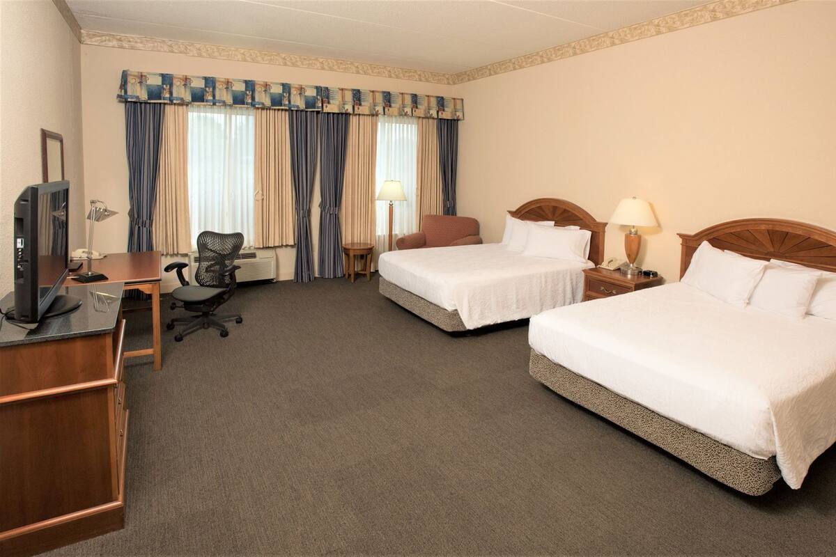 A hotel room with two beds, a desk, a chair, a TV, and a window with curtains.