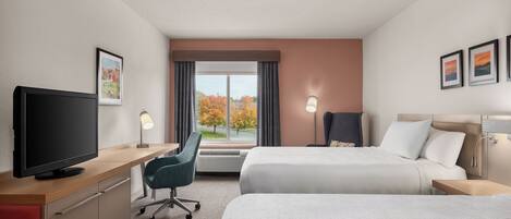 Habitación de hotel con dos camas, un escritorio con televisor, una silla y una ventana con vista a árboles.