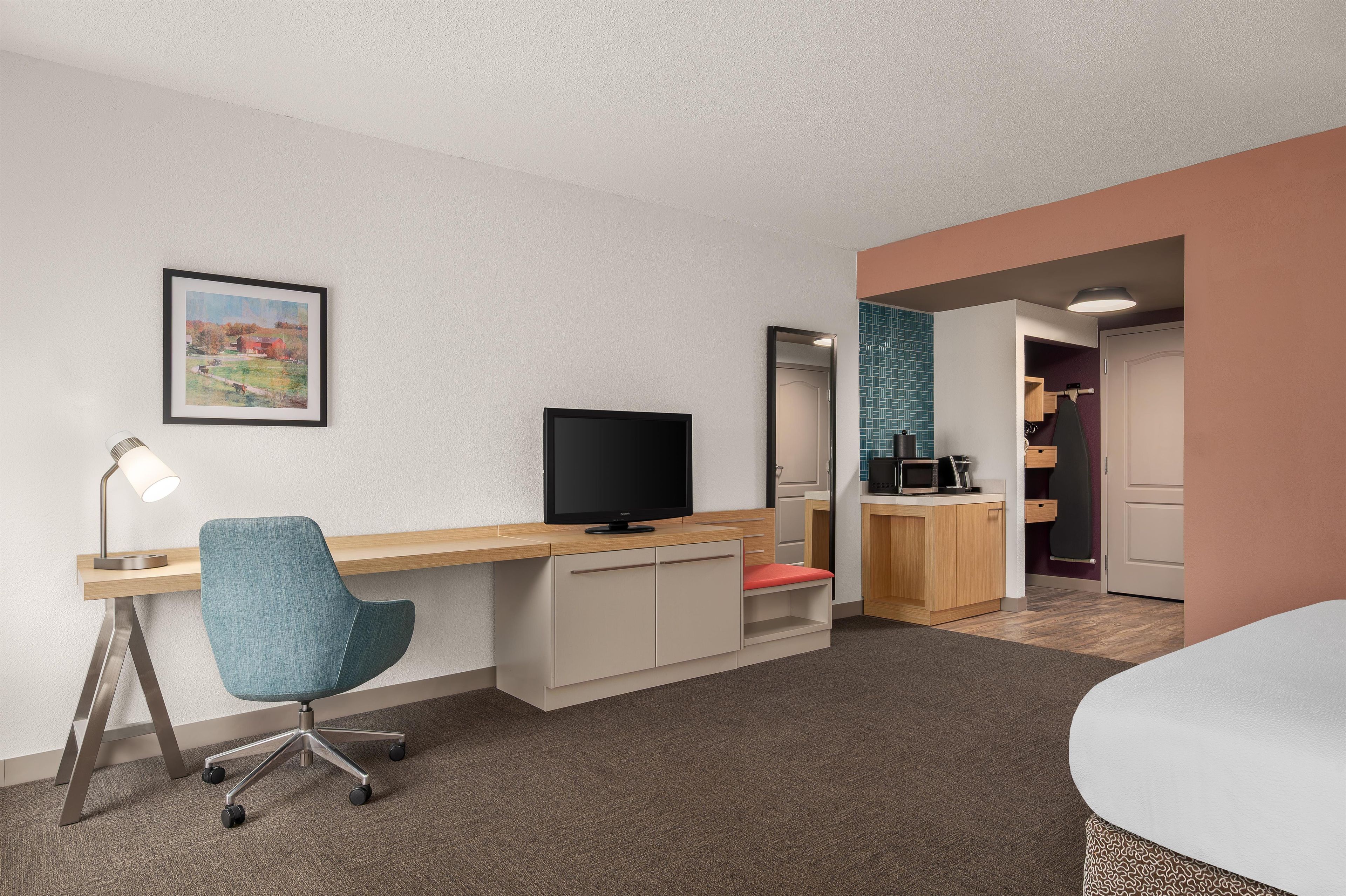 Una habitación de hotel con escritorio, silla, televisión y una kitchenette pequeña.