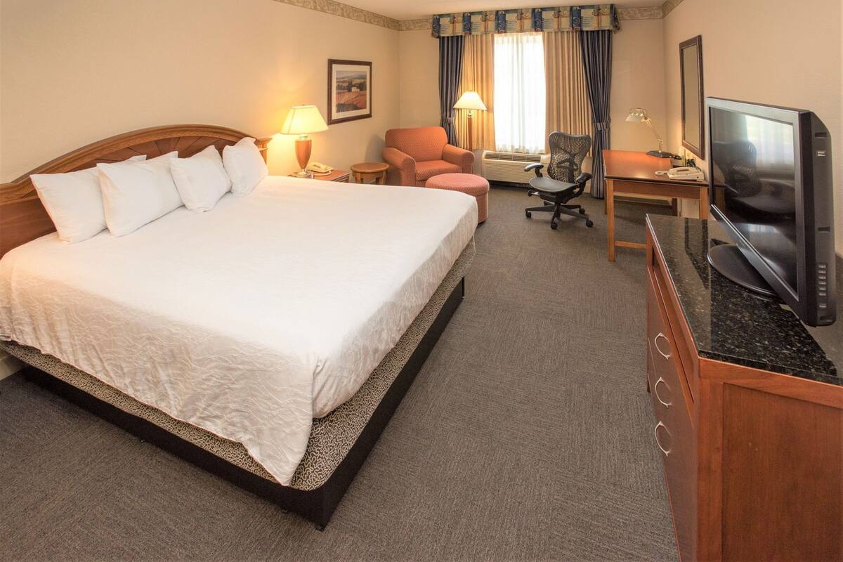 A hotel room with a large bed, a desk with a television, a chair, and a small table with a lamp.