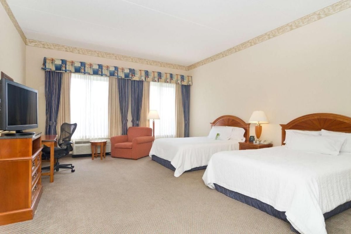 A hotel room with two beds, a television, a desk, a chair, and a window with curtains.