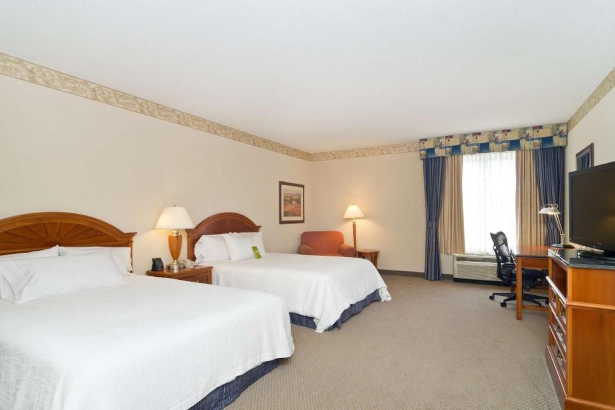 A hotel room with two beds, a desk, a chair, a television, and a window with curtains.