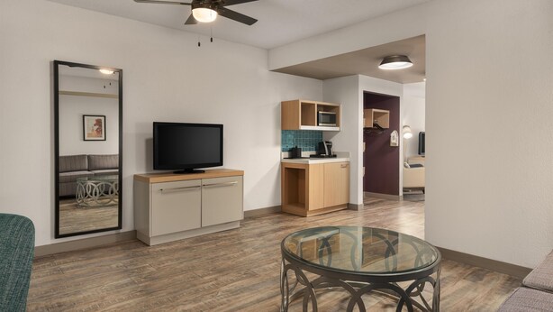 A modern living room with a flat-screen TV, a glass coffee table, a ceiling fan, and a kitchen area with wooden cabinets and a microwave. - Hilton Garden Inn Wooster (Wooster)
