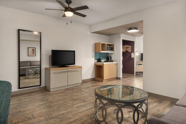 A modern living room with a flat-screen TV, a glass coffee table, a ceiling fan, and a kitchen area with wooden cabinets and a microwave.