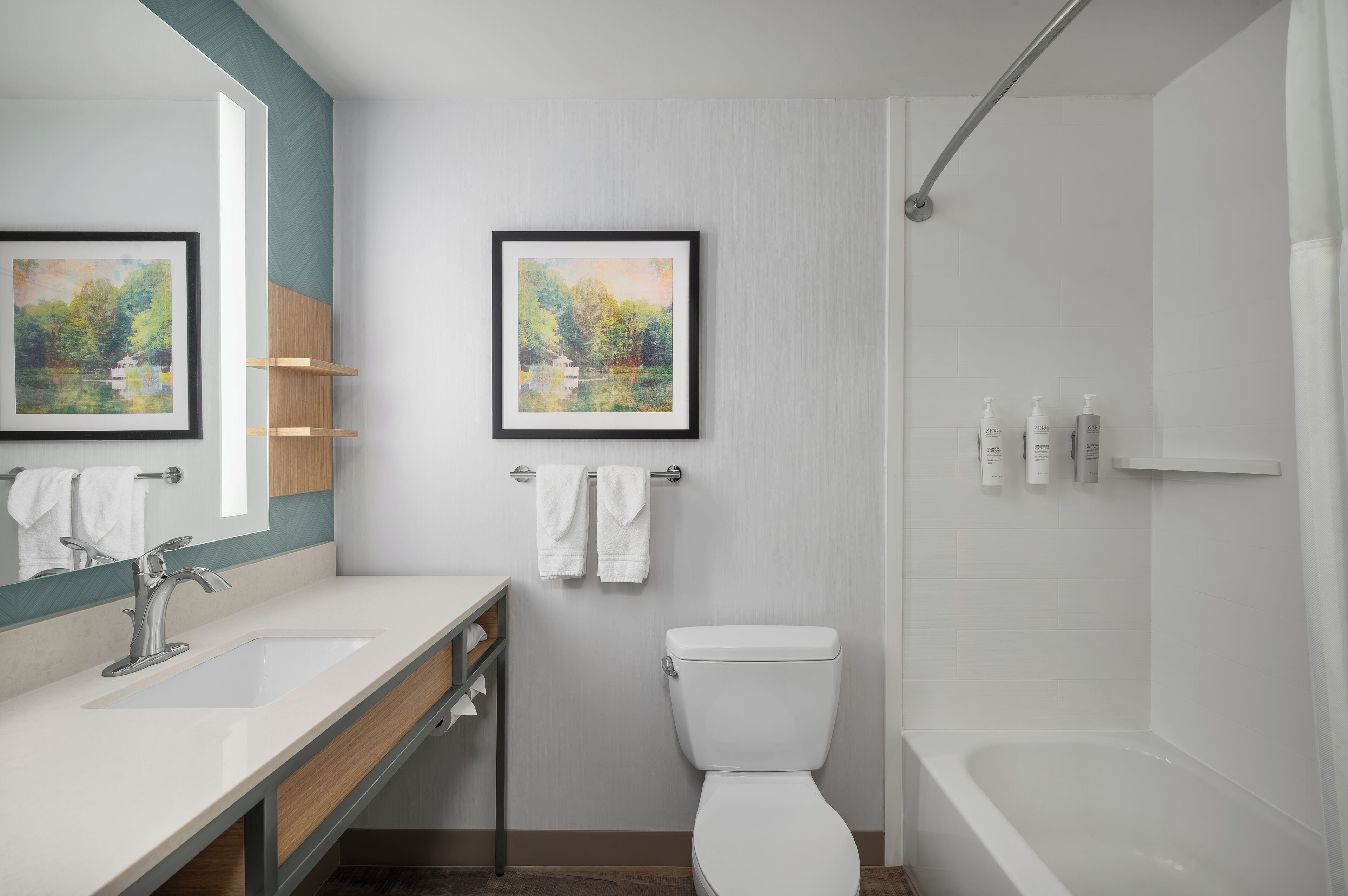 Un baño moderno con lavamanos, espejo, inodoro y ducha. Hay dos cuadros enmarcados en la pared y un perchero con toallas.