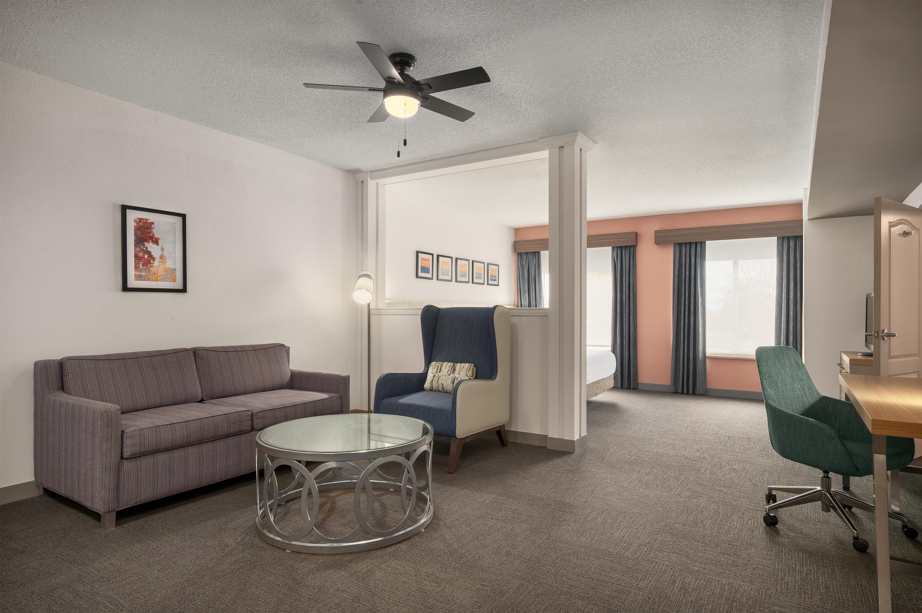 Habitación de hotel con sofá, una silla, una mesa redonda de vidrio, ventilador de techo y una cama con cortinas.
