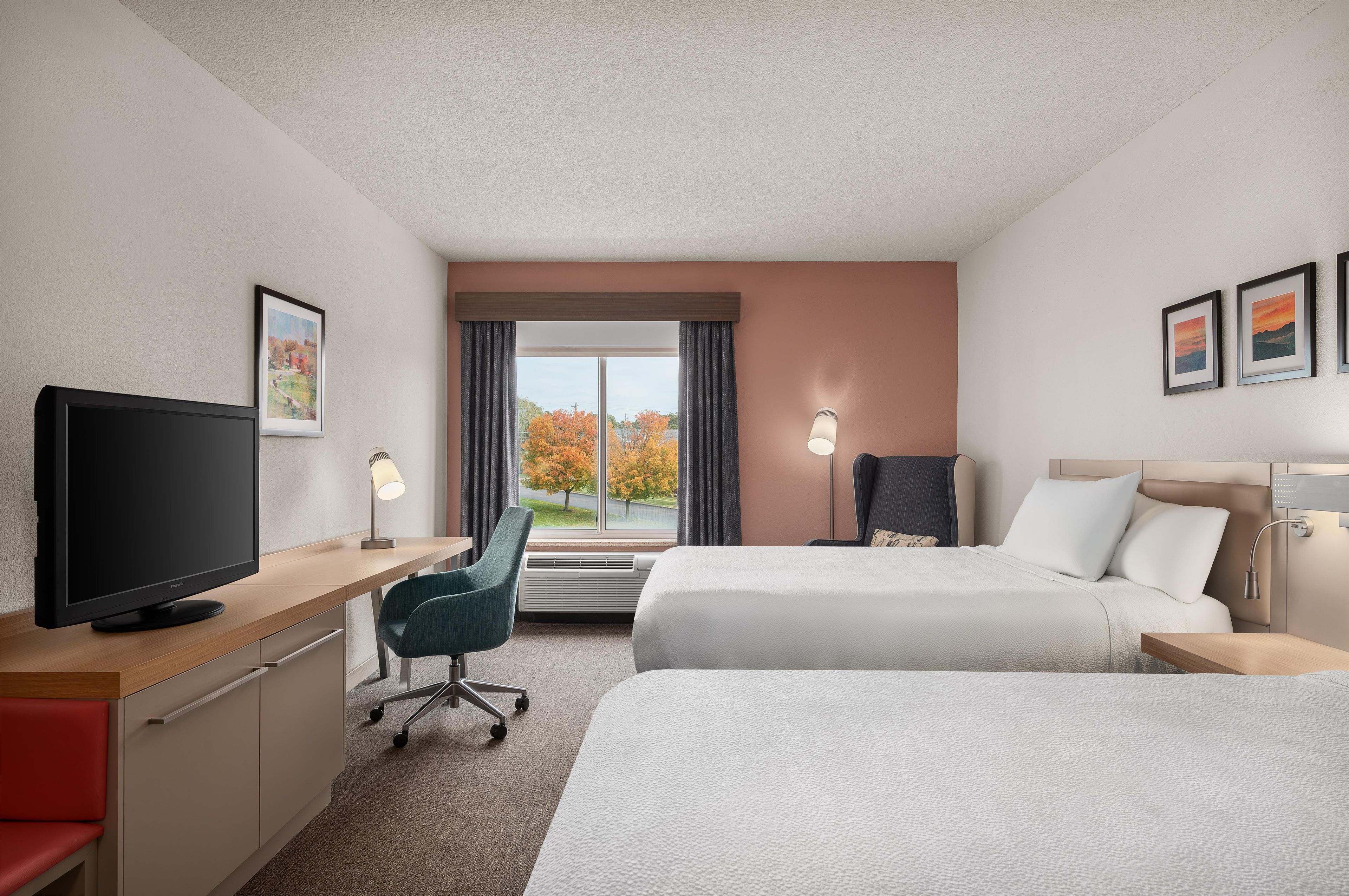 Habitación de hotel con cama, escritorio, silla, televisión y ventana con vista a árboles.