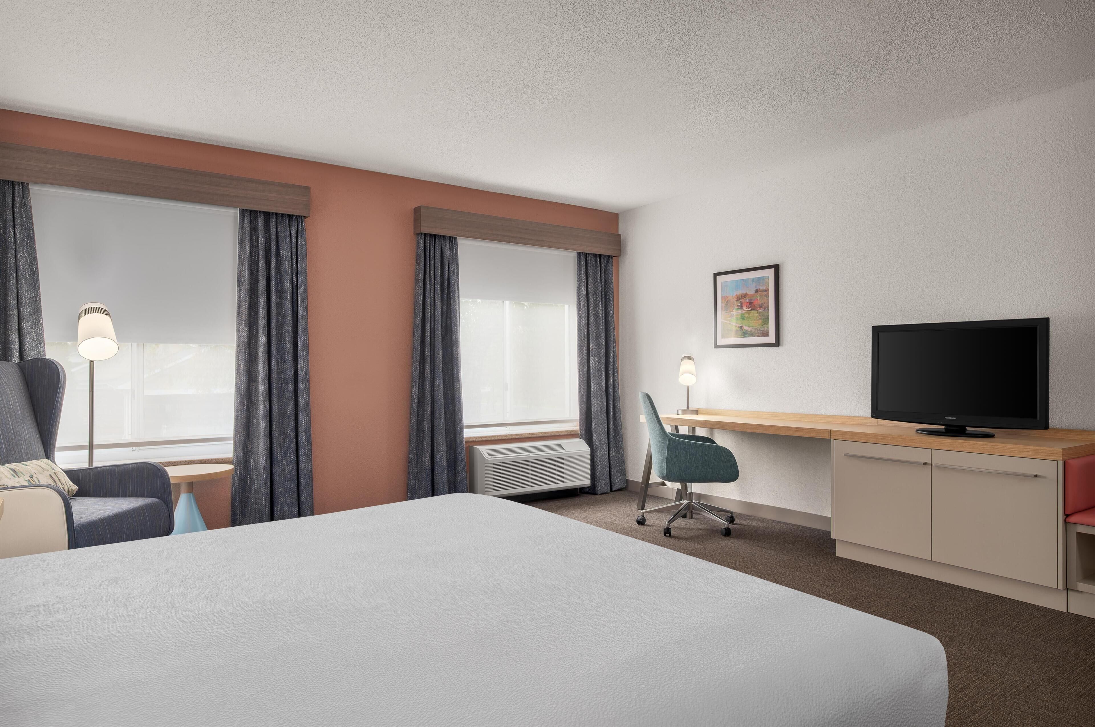 Une chambre d’hôtel avec un lit, un bureau avec une télévision, une chaise et une fenêtre avec des rideaux.