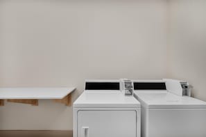 A laundry room with two white washing machines and a wooden shelf.