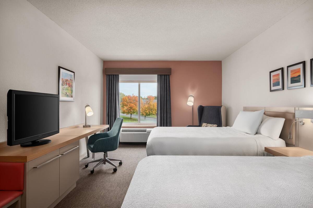 A hotel room with two beds, a desk with a TV, a chair, and a window with a view of trees.
