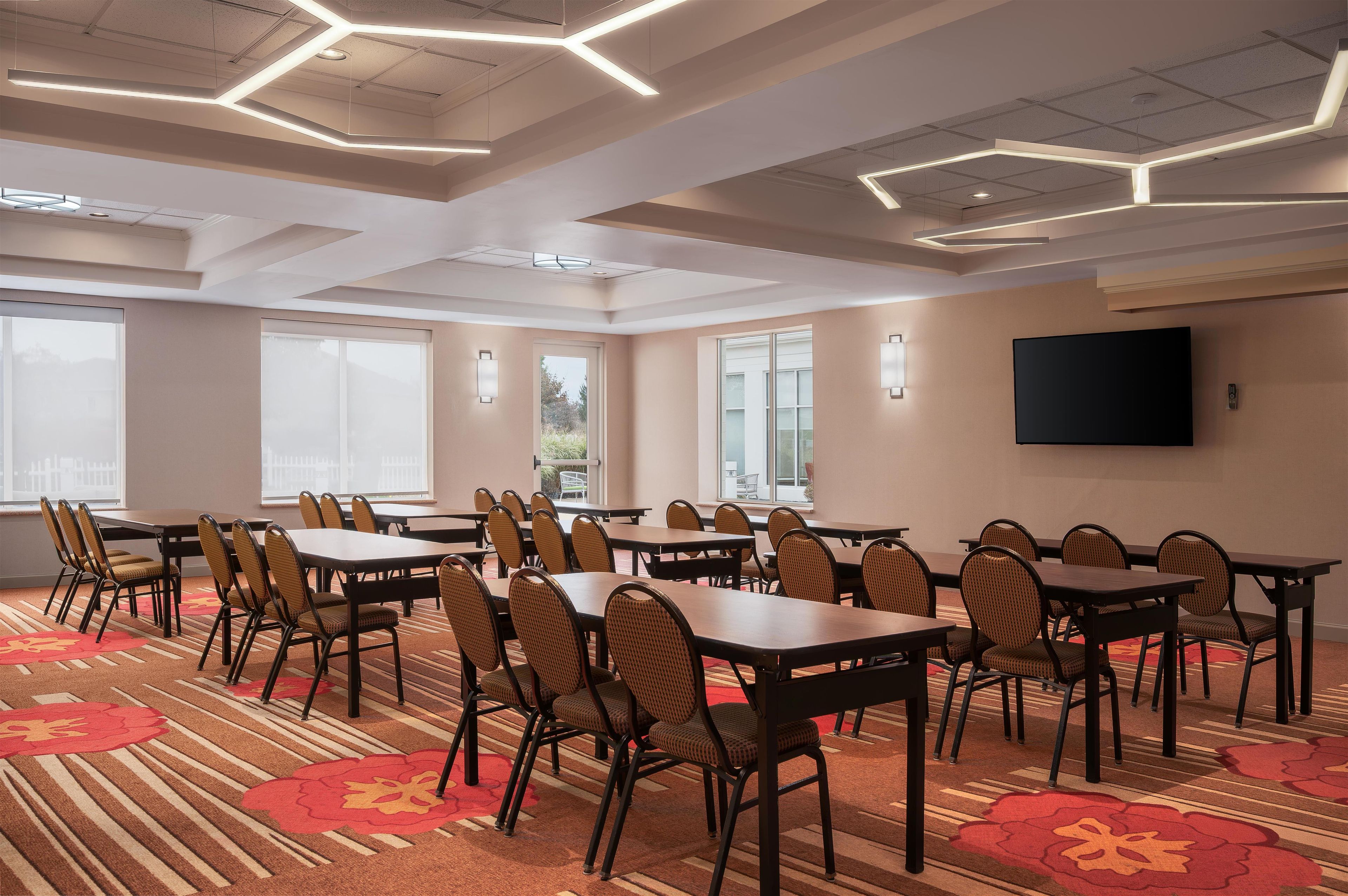 A conference room with tables and chairs, large windows, and a flat-screen TV on the wall.