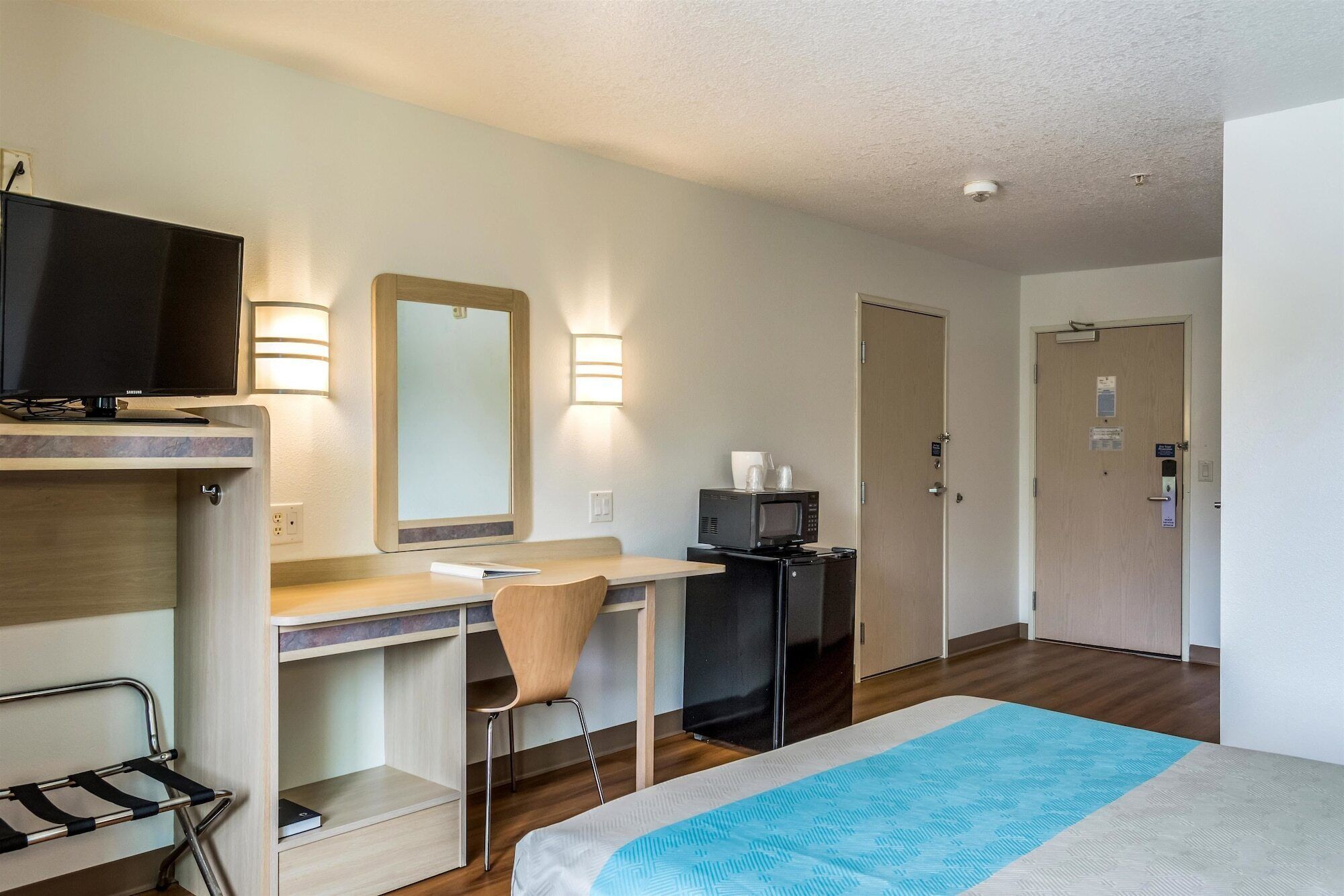 Habitación de hotel con cama, escritorio con silla, televisor, espejo, microondas y refrigerador.