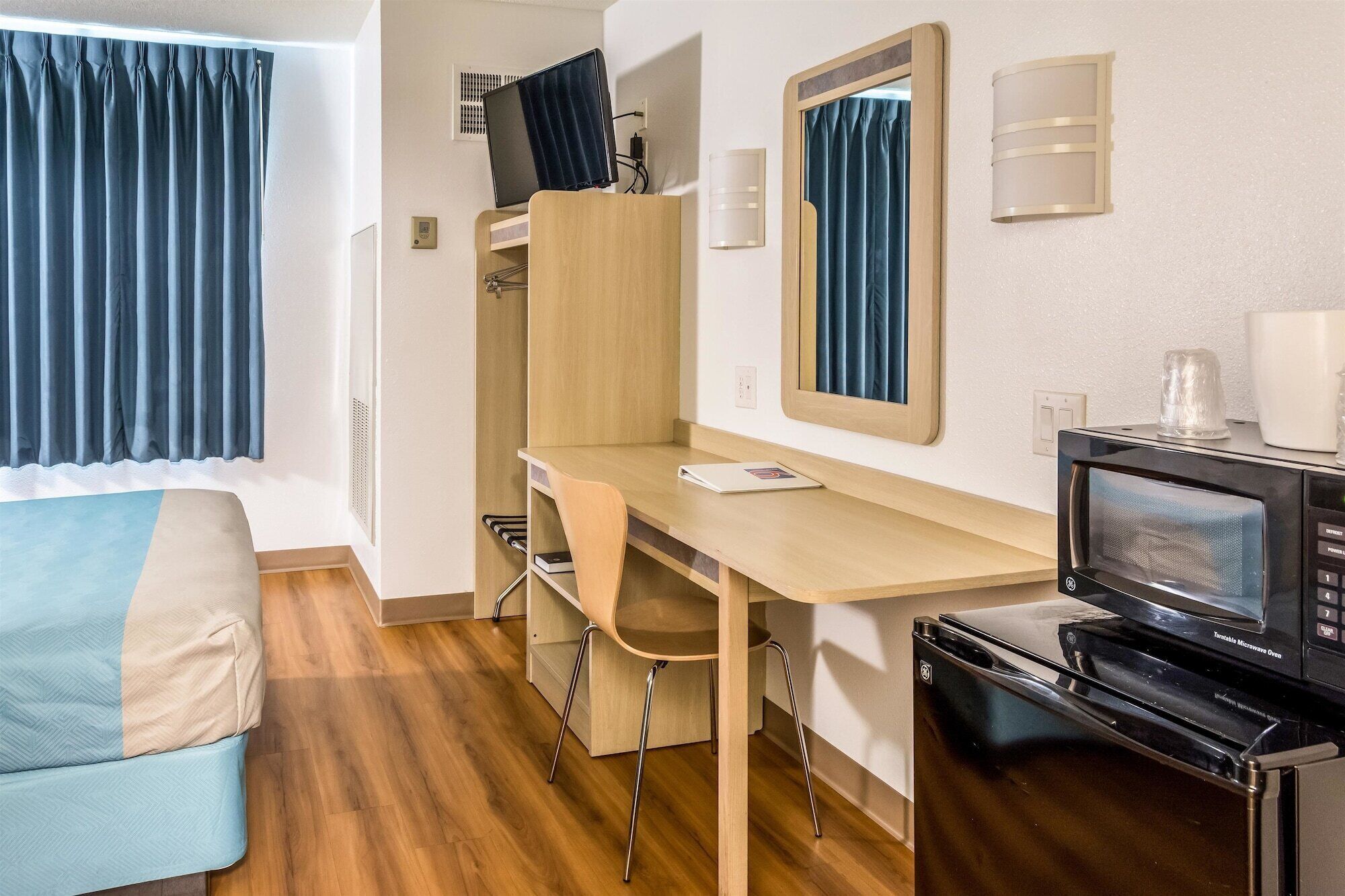 A compact room with a wooden desk, a chair, a bed with a blue and beige cover, a microwave oven, and a small kitchenette area.