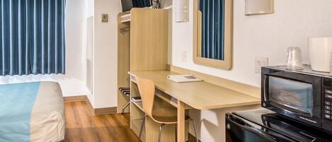 A compact room with a wooden desk, a chair, a bed with a blue and beige cover, a microwave oven, and a small kitchenette area.