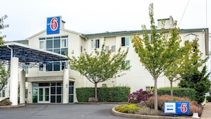 Edificio de Motel 6 con un letrero azul, entrada cubierta y un área ajardinada con árboles y arbustos.