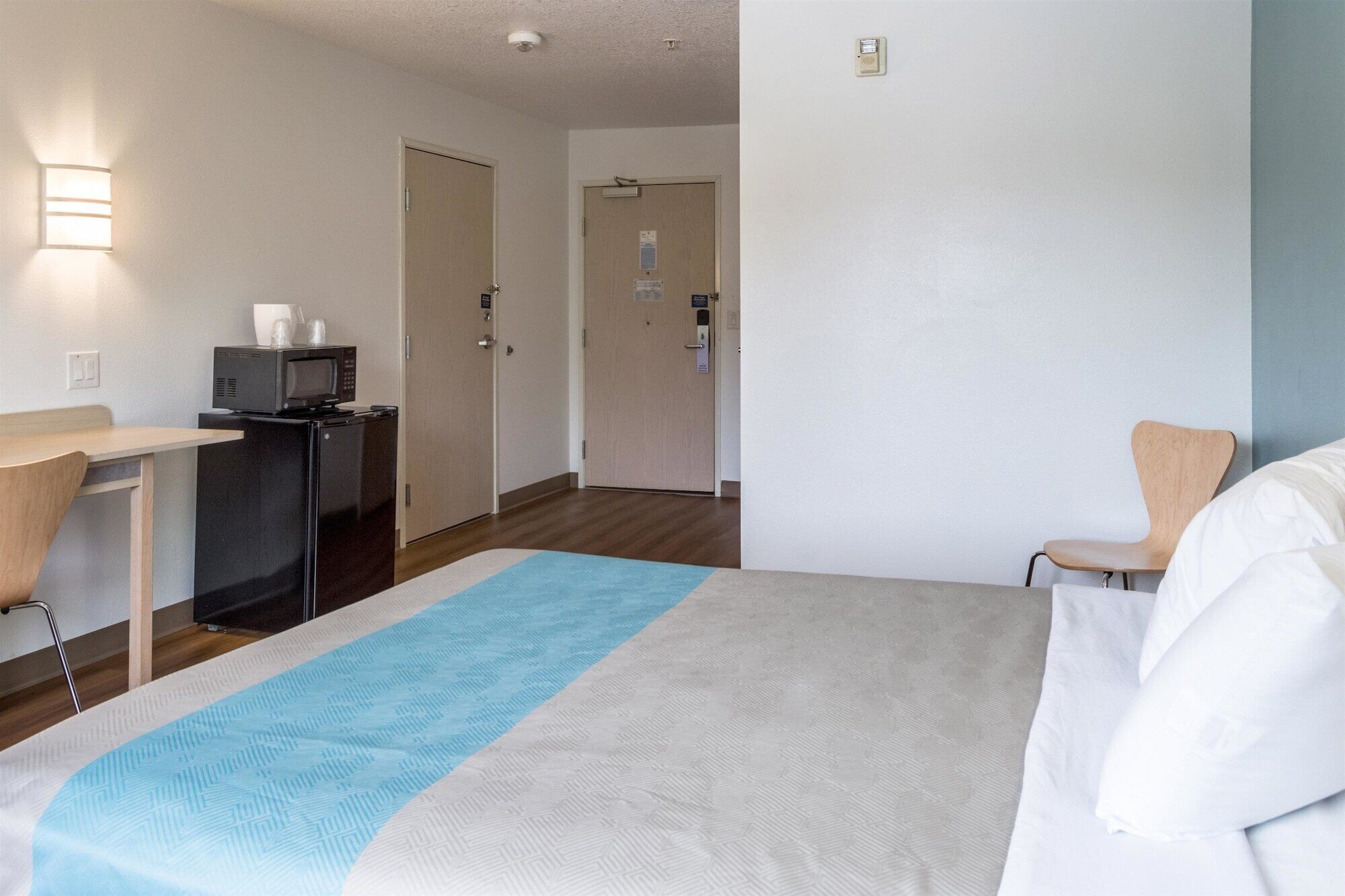 A hotel room with a bed, a desk with a microwave, a chair, and a door leading to another room.