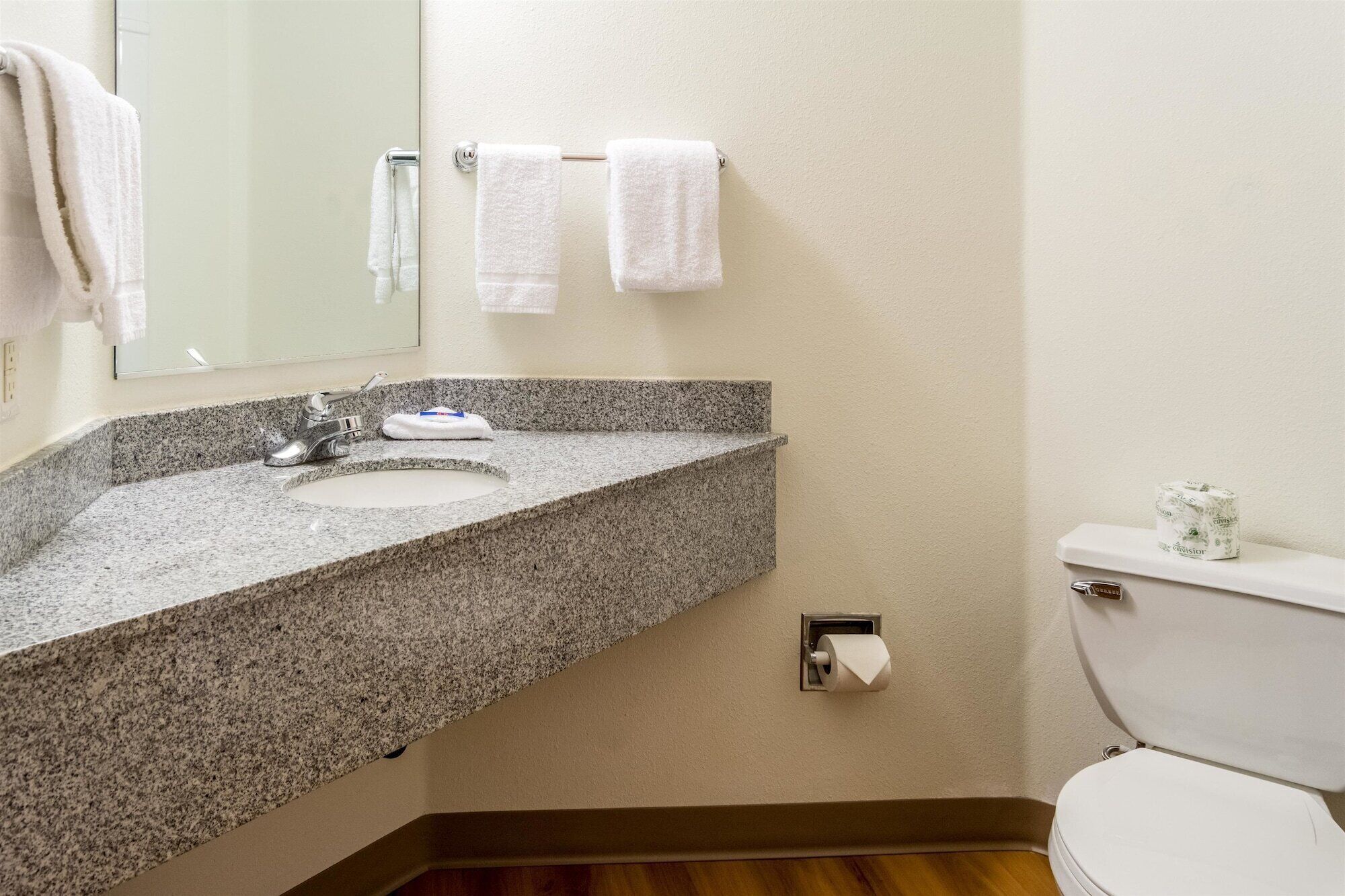 Un baño con lavamanos de encimera de granito, un espejo, dos toallas colgadas en un perchero y un inodoro con dispensador de papel higiénico.