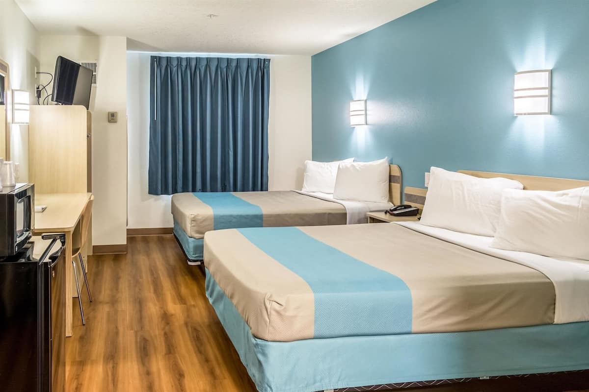 Een hotelkamer met twee bedden, een bureau met een computer, een telefoon en een raam met blauwe gordijnen.