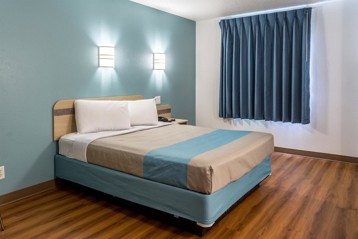 Een hotelkamer met een bed, een houten hoofdbord, nachtlampjes, een raam met gordijnen en een houten vloer.