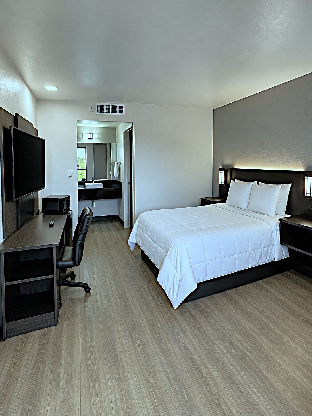 A hotel room with a bed, a desk with a chair, a television, and a bathroom visible through an open door.