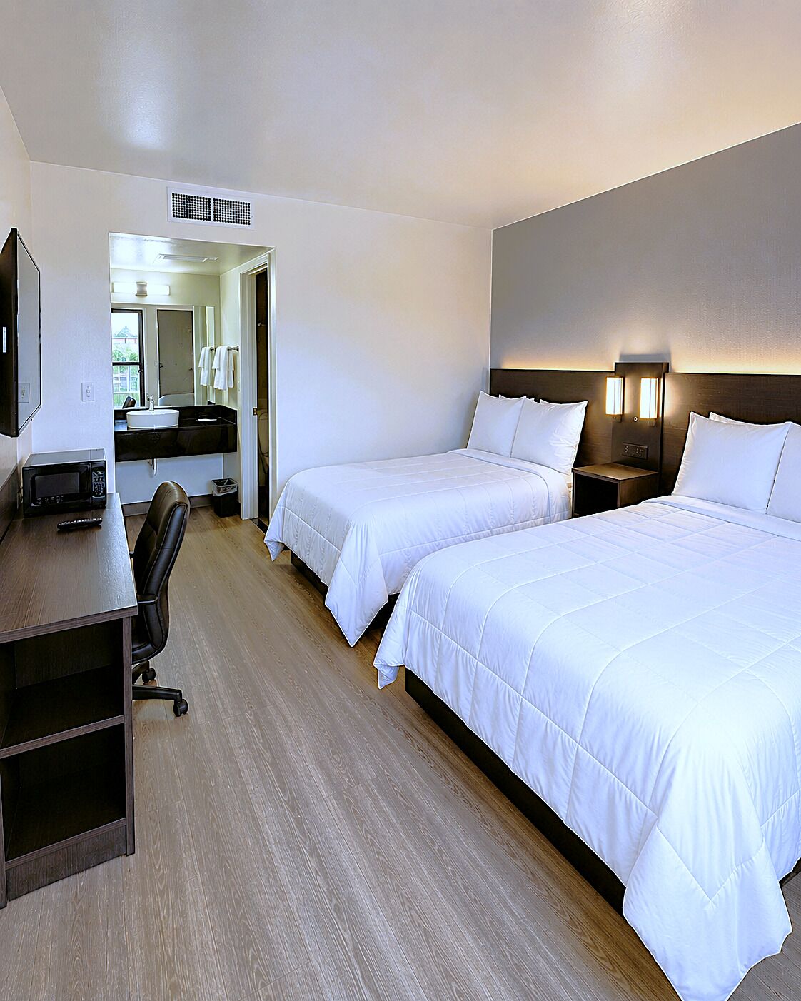 A hotel room with two beds, a desk, a chair, a microwave, a sink, and a mirror.