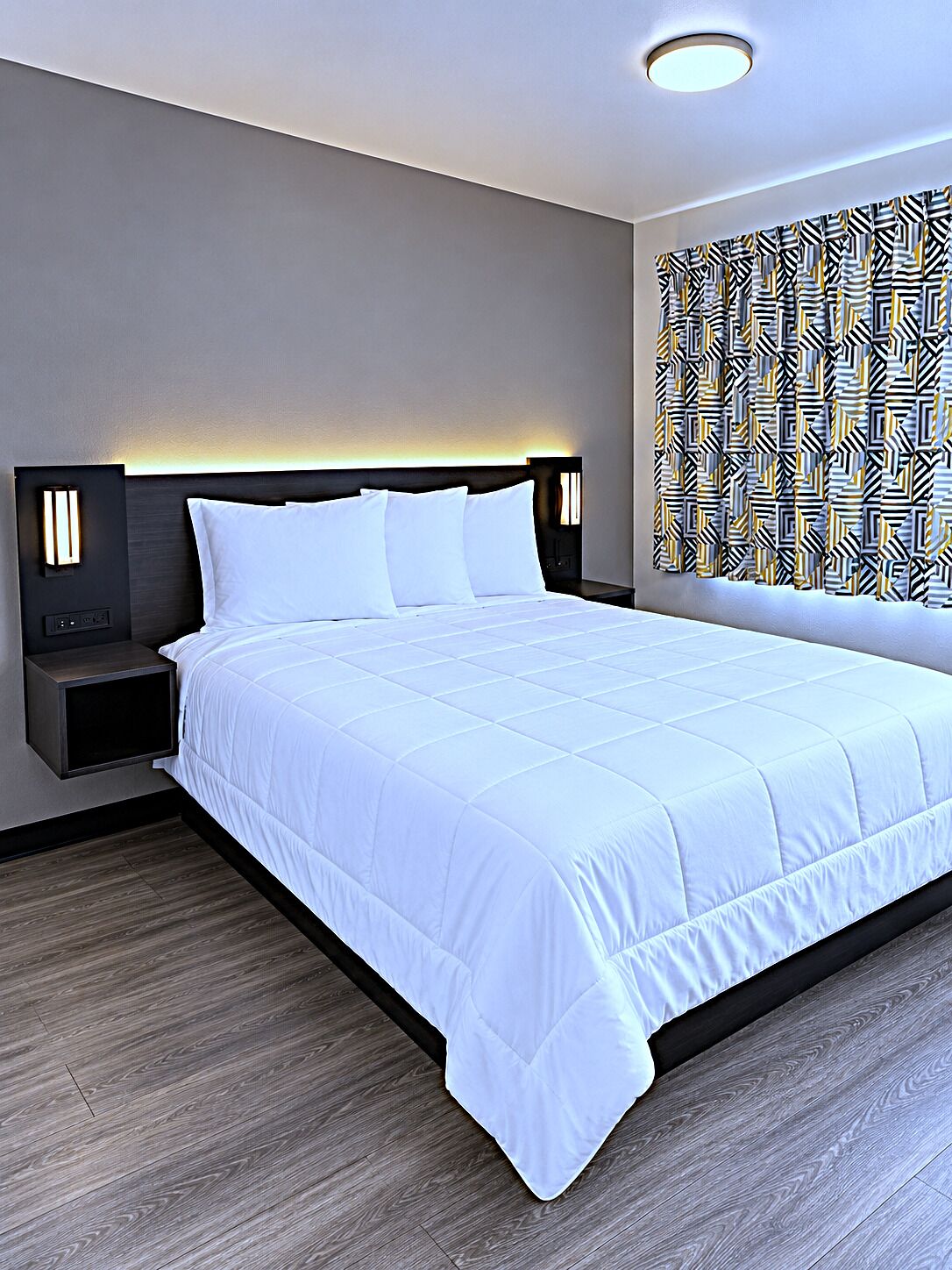 A hotel room with a large bed, bedside lamps, a patterned curtain, and a modern ceiling light.