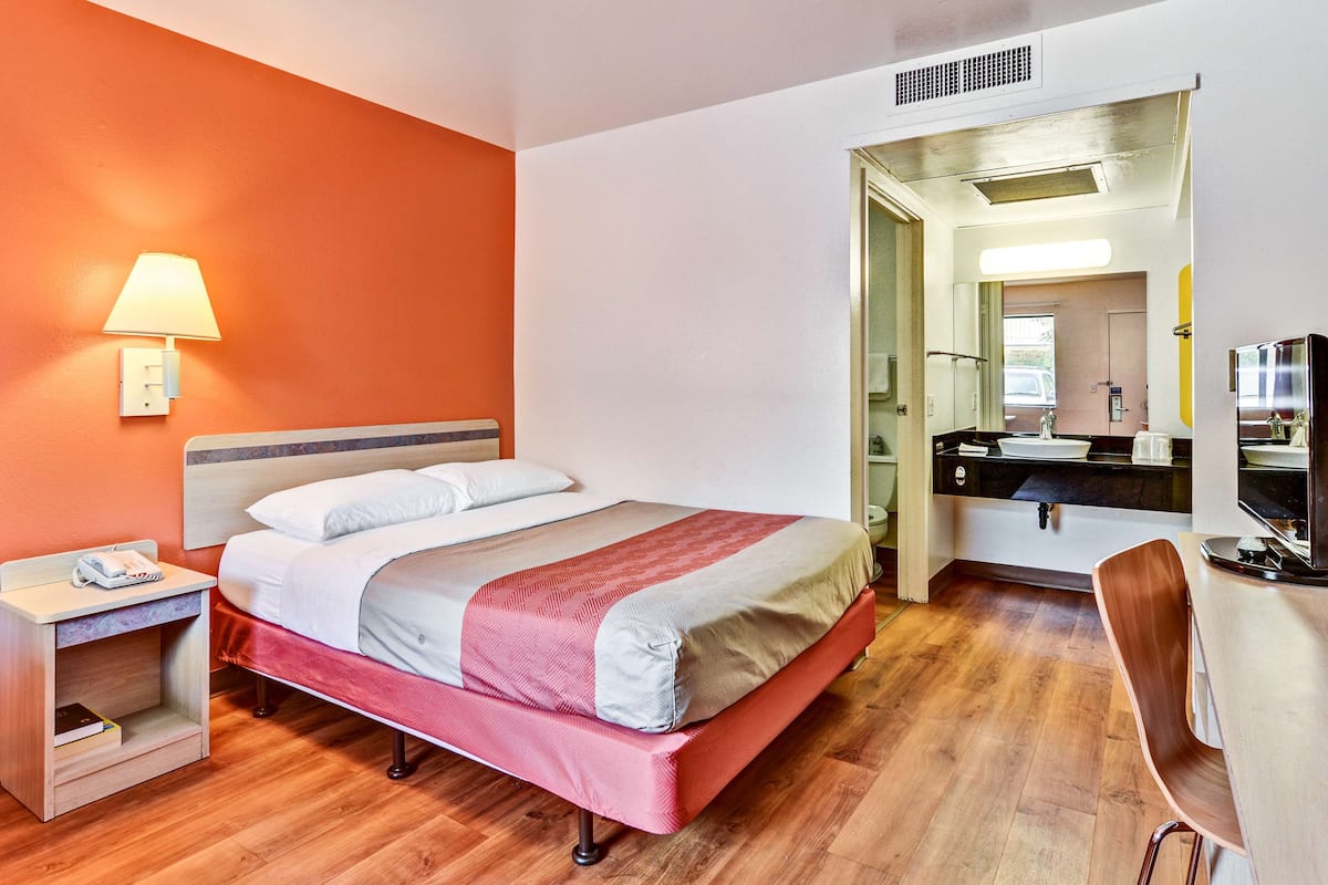 A hotel room with a bed, a desk, a chair, a lamp, and a kitchenette.