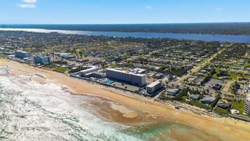 An aerial view of a coastal town with a beachfront hotel, residential areas, and a large body of water in the background.
