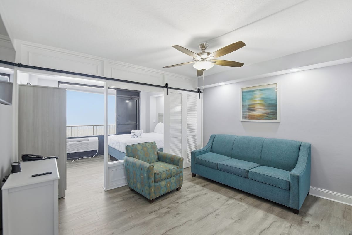 Une chambre d’hôtel moderne avec un balcon, un canapé bleu, un fauteuil à motifs et un ventilateur de plafond.