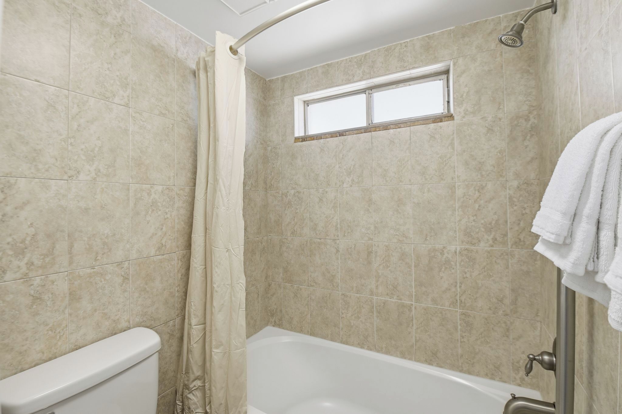 Un baño con baldosas color beige, una bañera, una ducha con cortina y una ventana sobre la ducha.