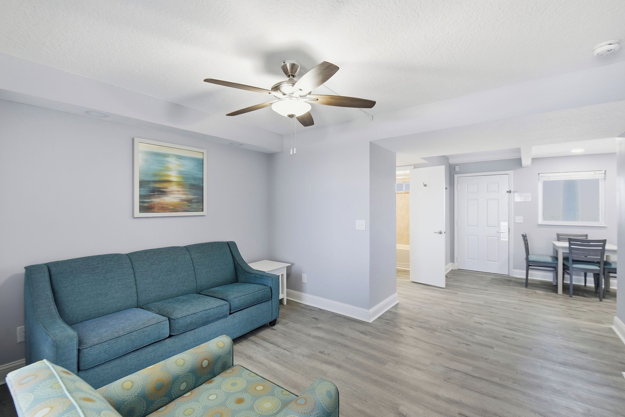 A living room with a blue sofa, a patterned armchair, a ceiling fan, and a painting on the wall.