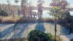 A tennis court with a surrounding fence, a palm tree, and a body of water in the background. - Lifetime of Vacations Resort (Kissimmee)