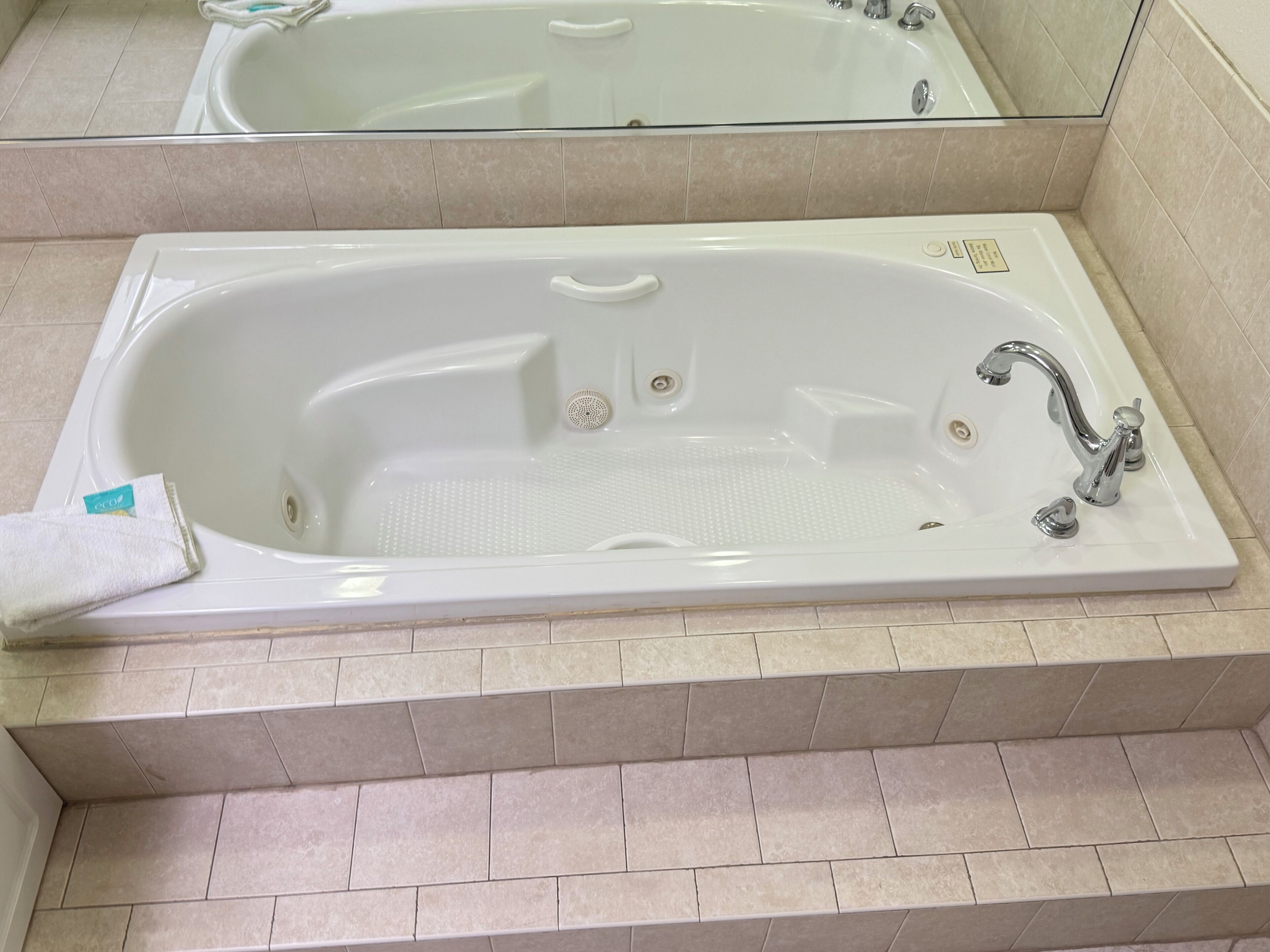 Una bañera blanca con dos grifos y un desagüe, ubicada en un baño con suelo y paredes de azulejos color beige.