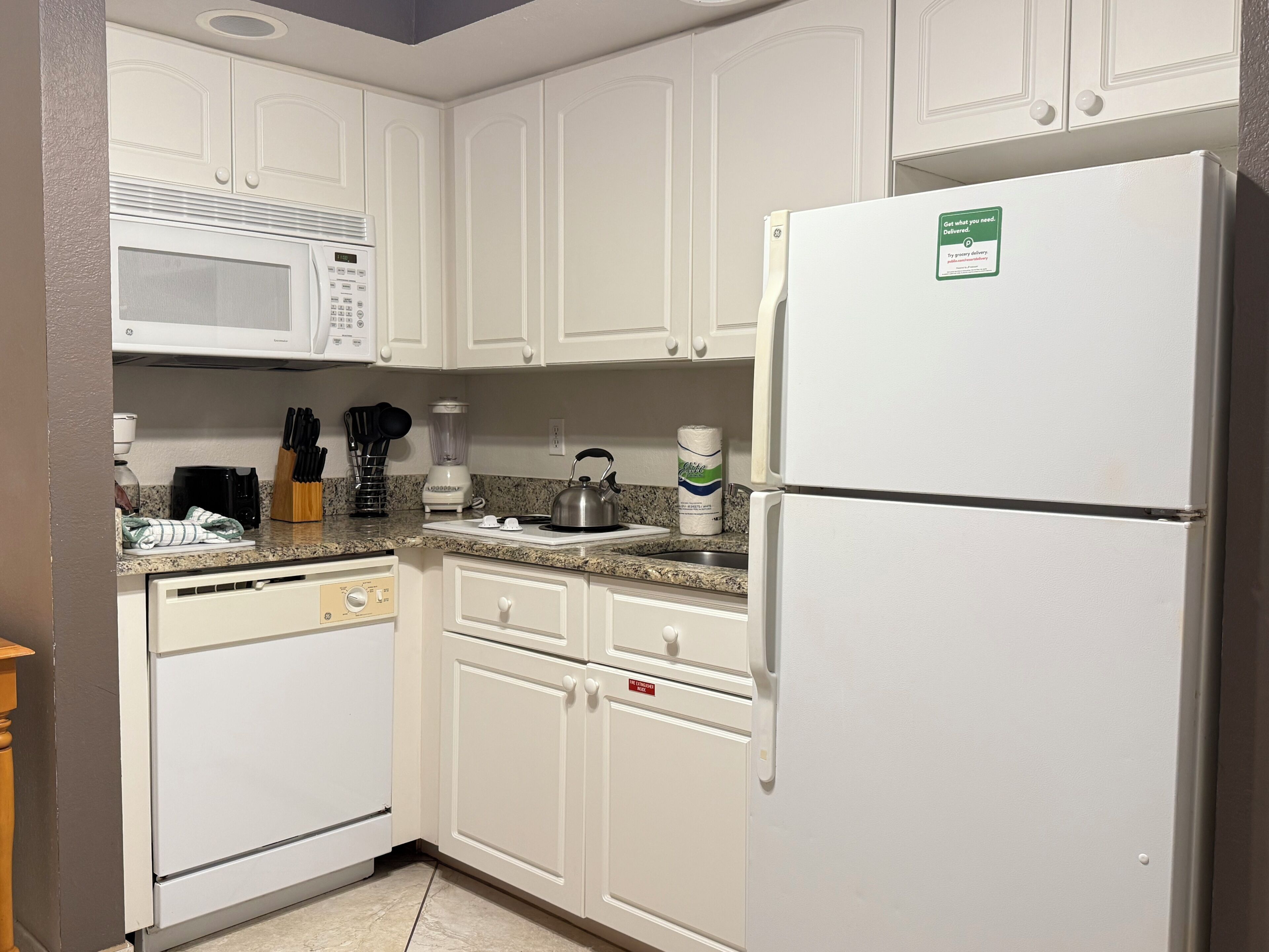 A kitchen with white cabinets, a microwave, a refrigerator, a kettle, and various kitchen utensils.