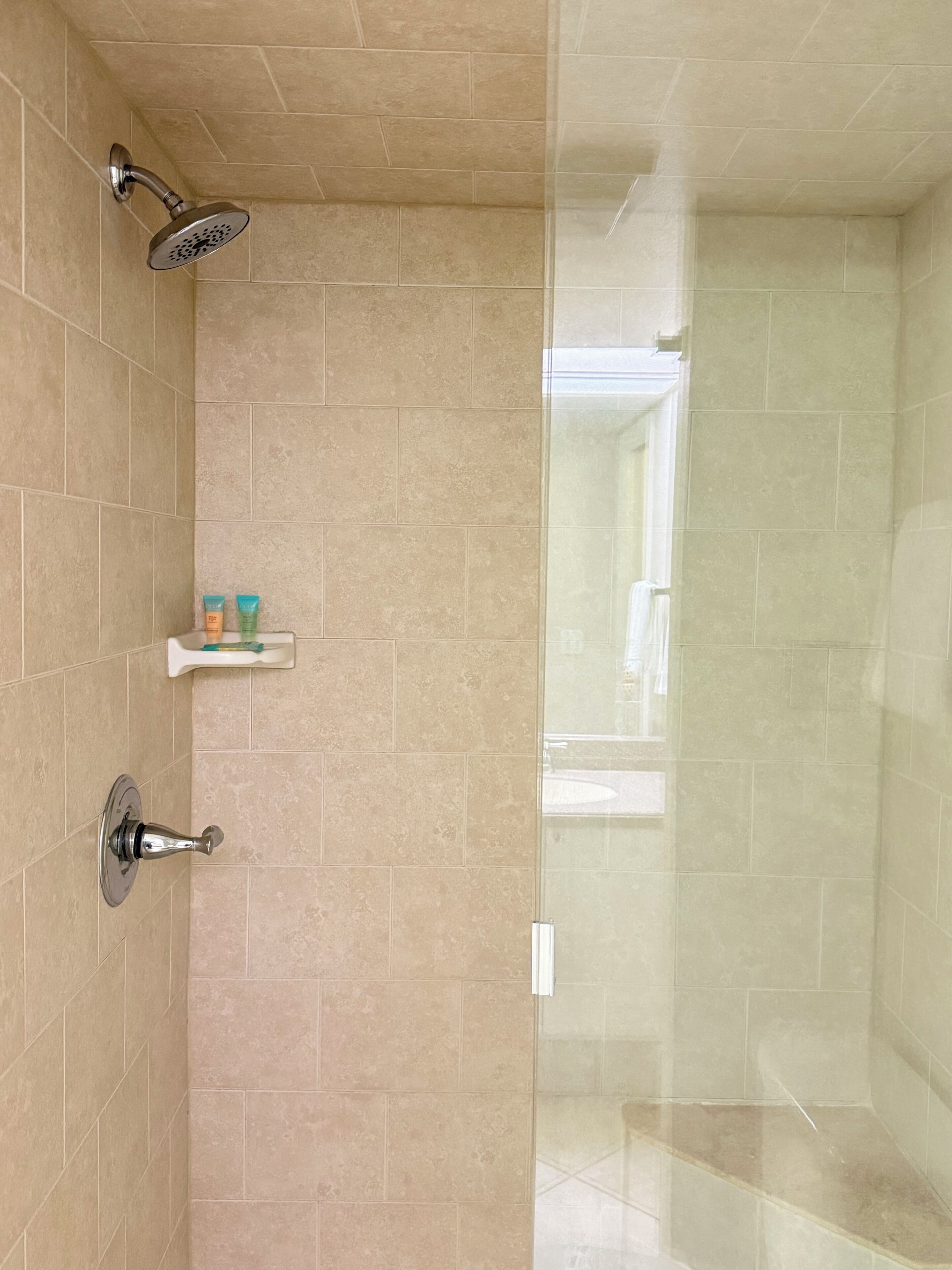 Zona de ducha con azulejos color beige, una puerta de vidrio y un pequeño estante con artículos de aseo.