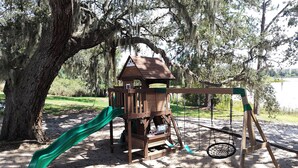 A wooden play structure with a slide and swings, surrounded by trees and sand. - Lifetime of Vacations Resort (Kissimmee)