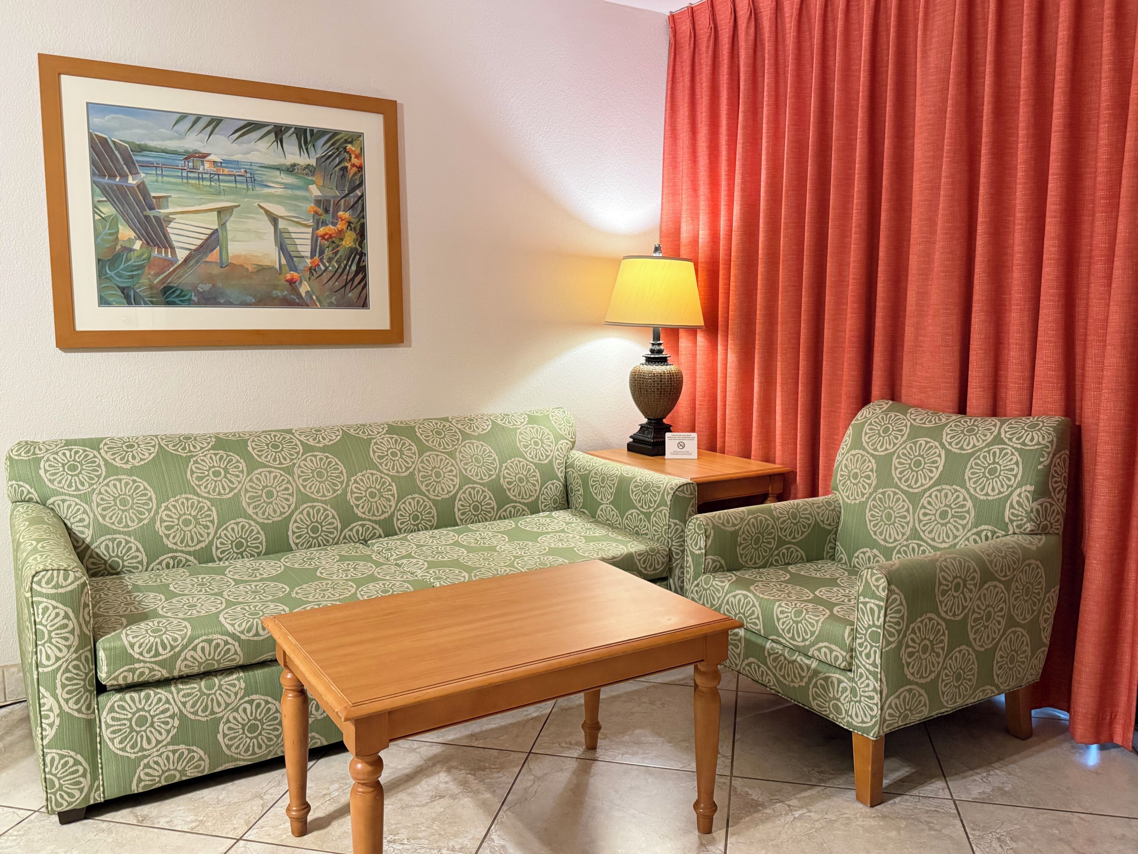 Una habitación con un sofá de color verde con estampado, una mesa de centro de madera, una lámpara y un cuadro de una escena tropical.