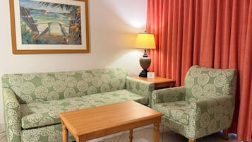A room with a green patterned sofa, a wooden coffee table, a lamp, and a painting of a tropical scene.