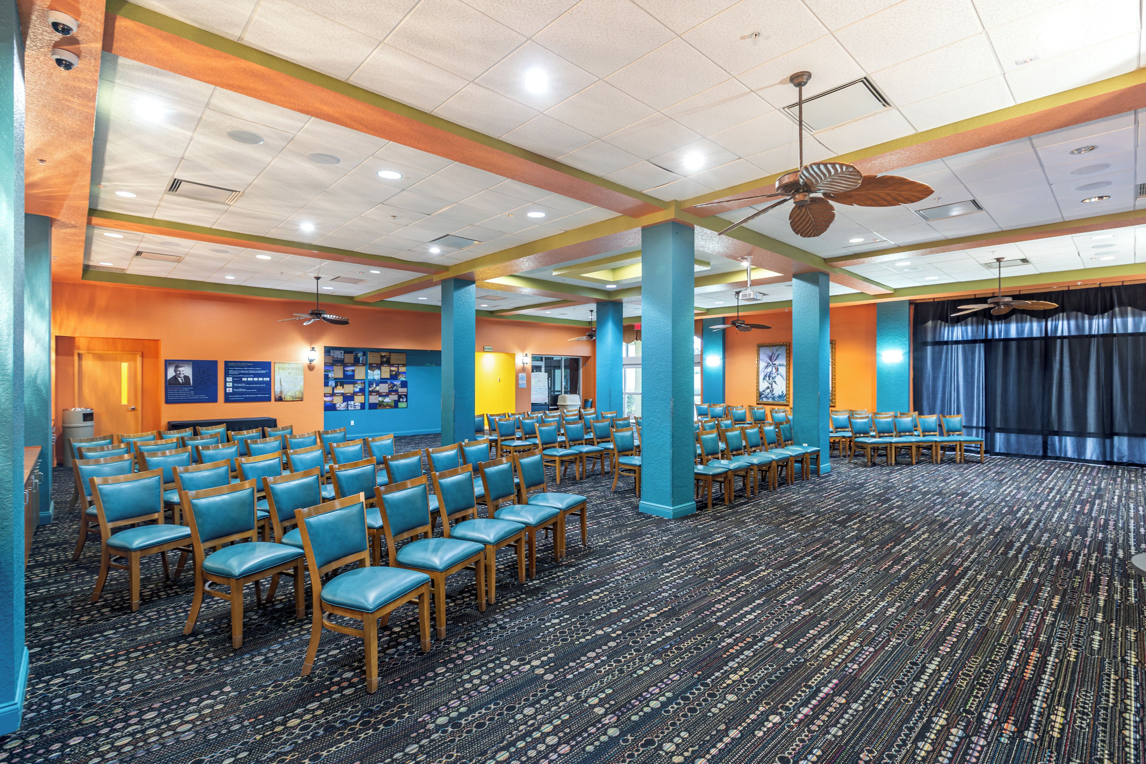 Un amplio salón de conferencias con filas de sillas, un escenario y ventiladores de techo.