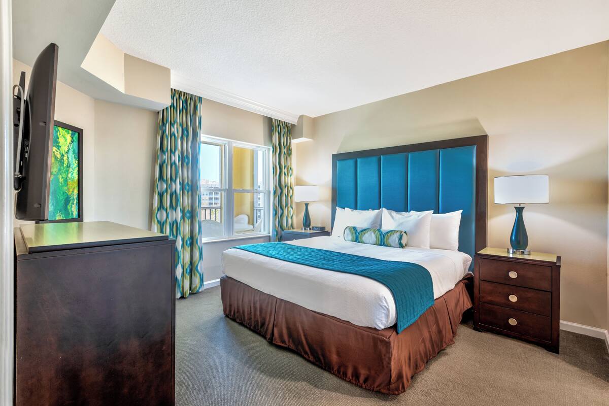 A hotel room with a large bed, a flat-screen TV, a desk, and a window with curtains.