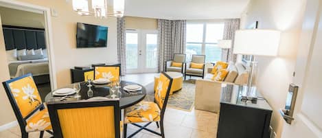 A hotel room with a dining area, a living room with a sofa and armchairs, a TV mounted on the wall, and a balcony with curtains.