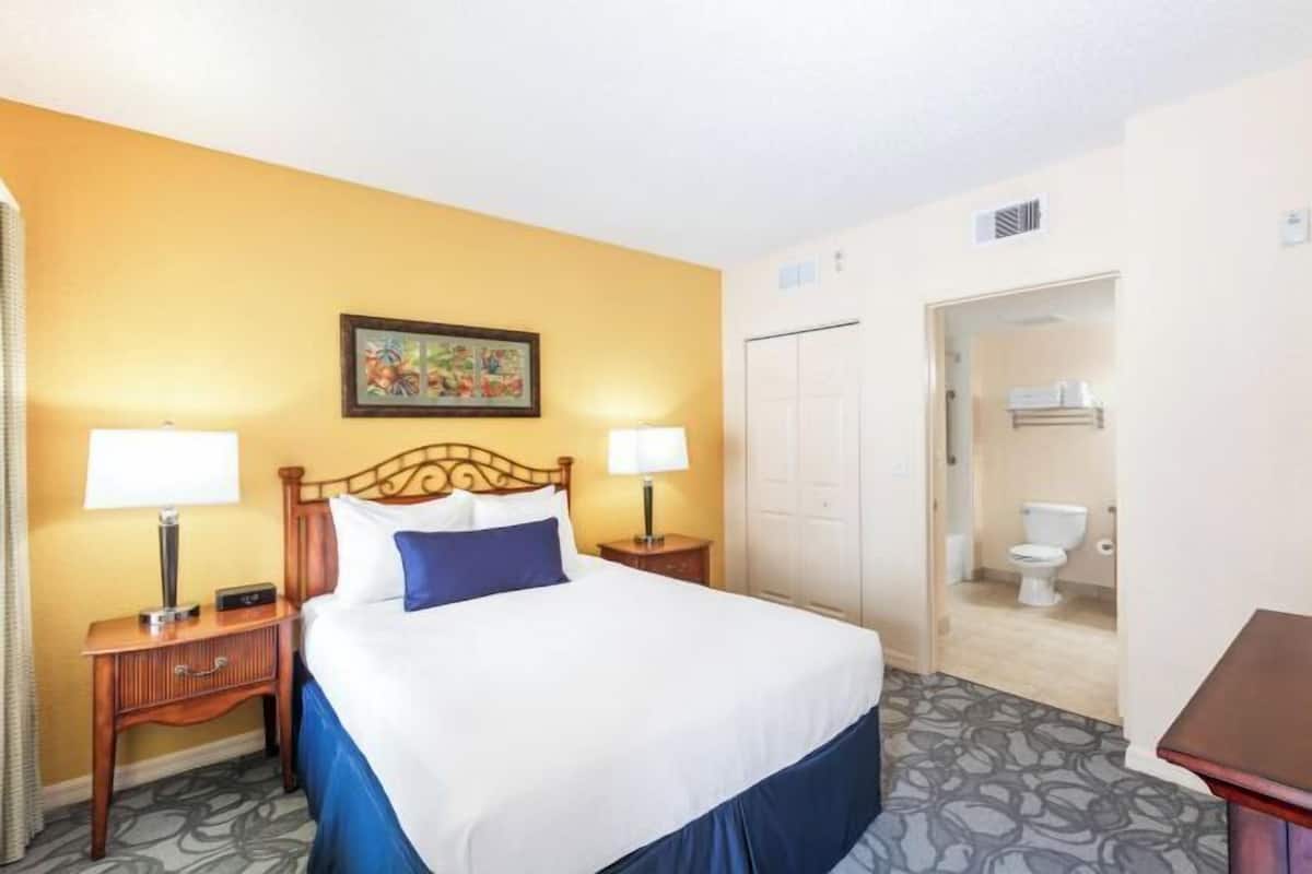A hotel room with a bed, two bedside tables, a nightstand, a painting on the wall, a door leading to a bathroom, and a small table.