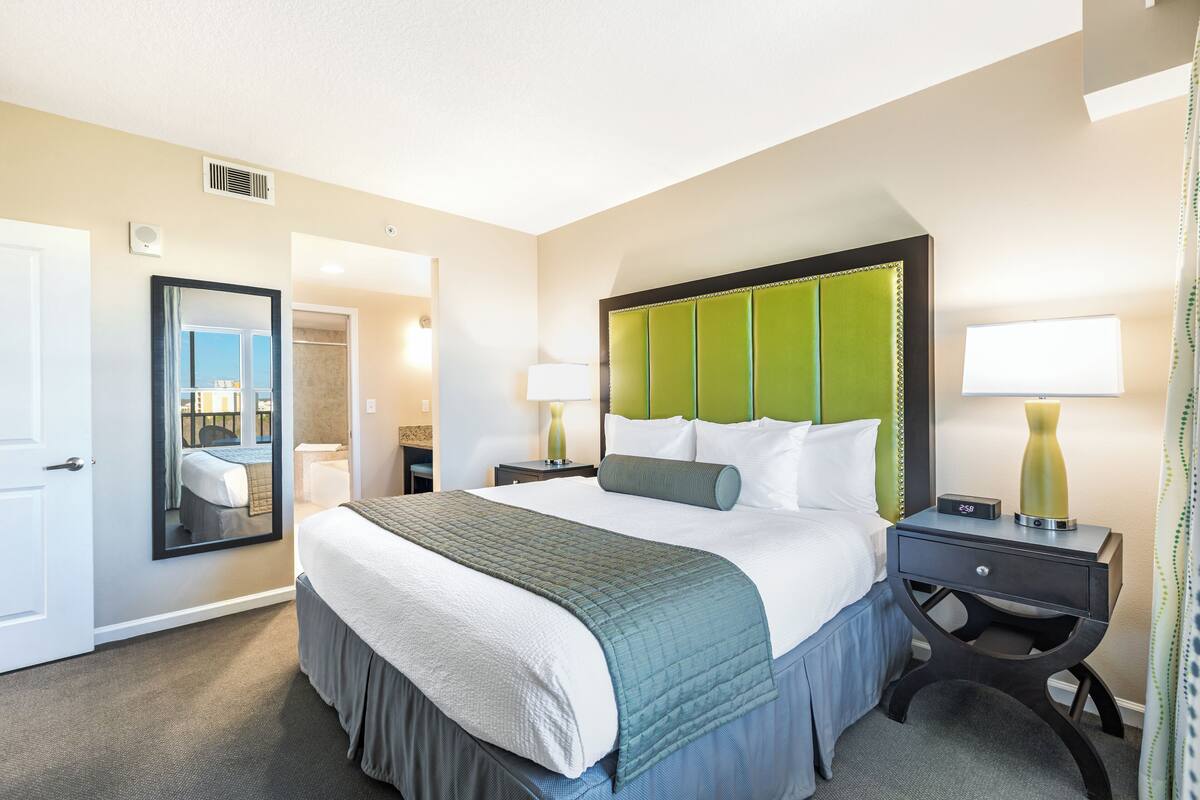 A hotel room with a large bed, a nightstand, a mirror, and a bathroom visible through an open door.