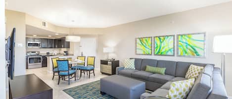 A modern living room with a grey sofa, blue and yellow chairs, a dining table, and a kitchen in the background.