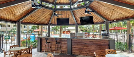 Un espace extérieur aménagé en terrasse, avec des tables et des chaises en bois, un comptoir de bar et des ventilateurs de plafond.