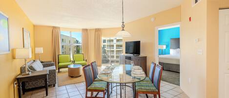 A hotel room with a dining area, a sofa, a TV, and a balcony with a view of other buildings.