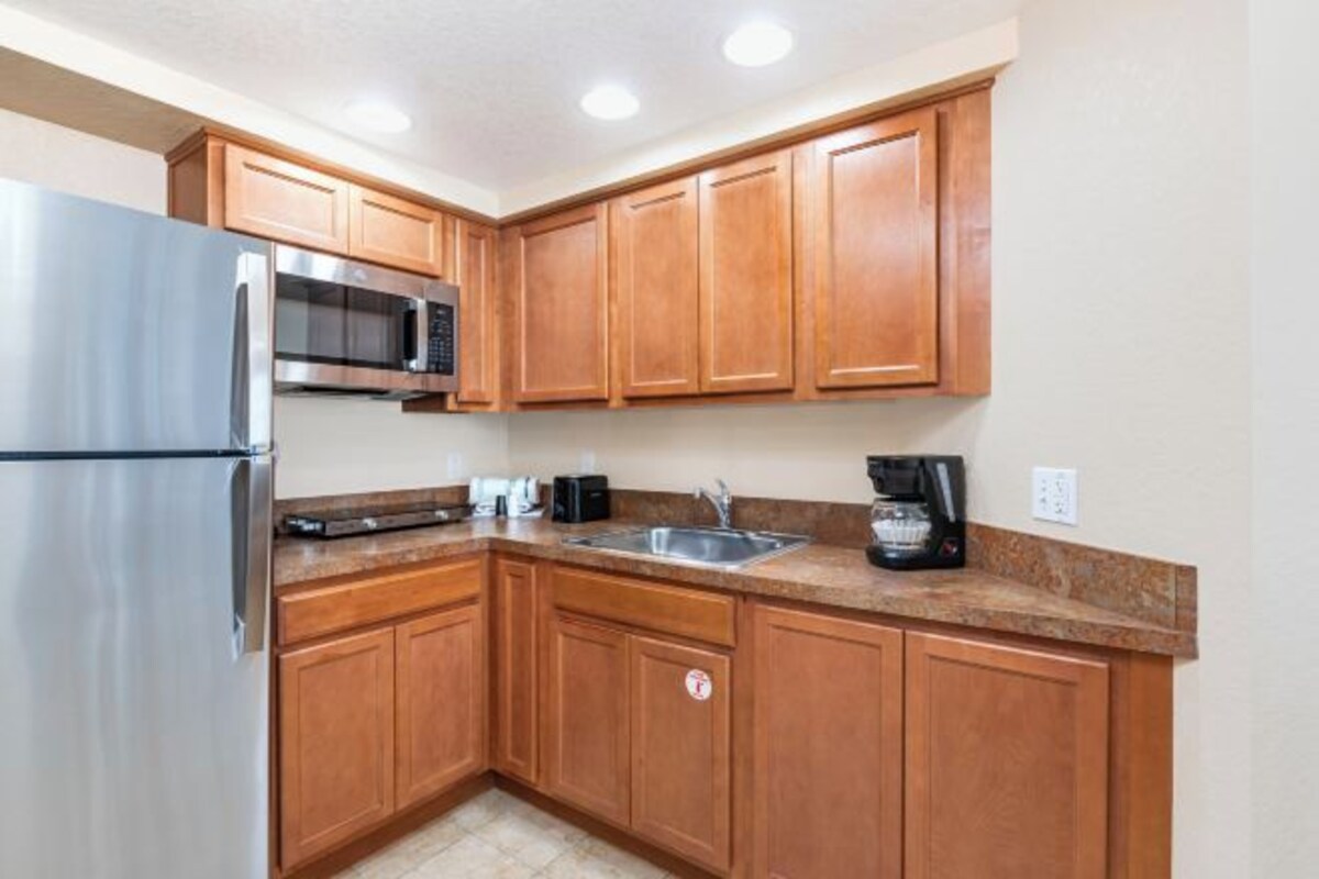 Una cocina con armarios de madera, un refrigerador de acero inoxidable, un microondas, un lavamanos y una cafetera.