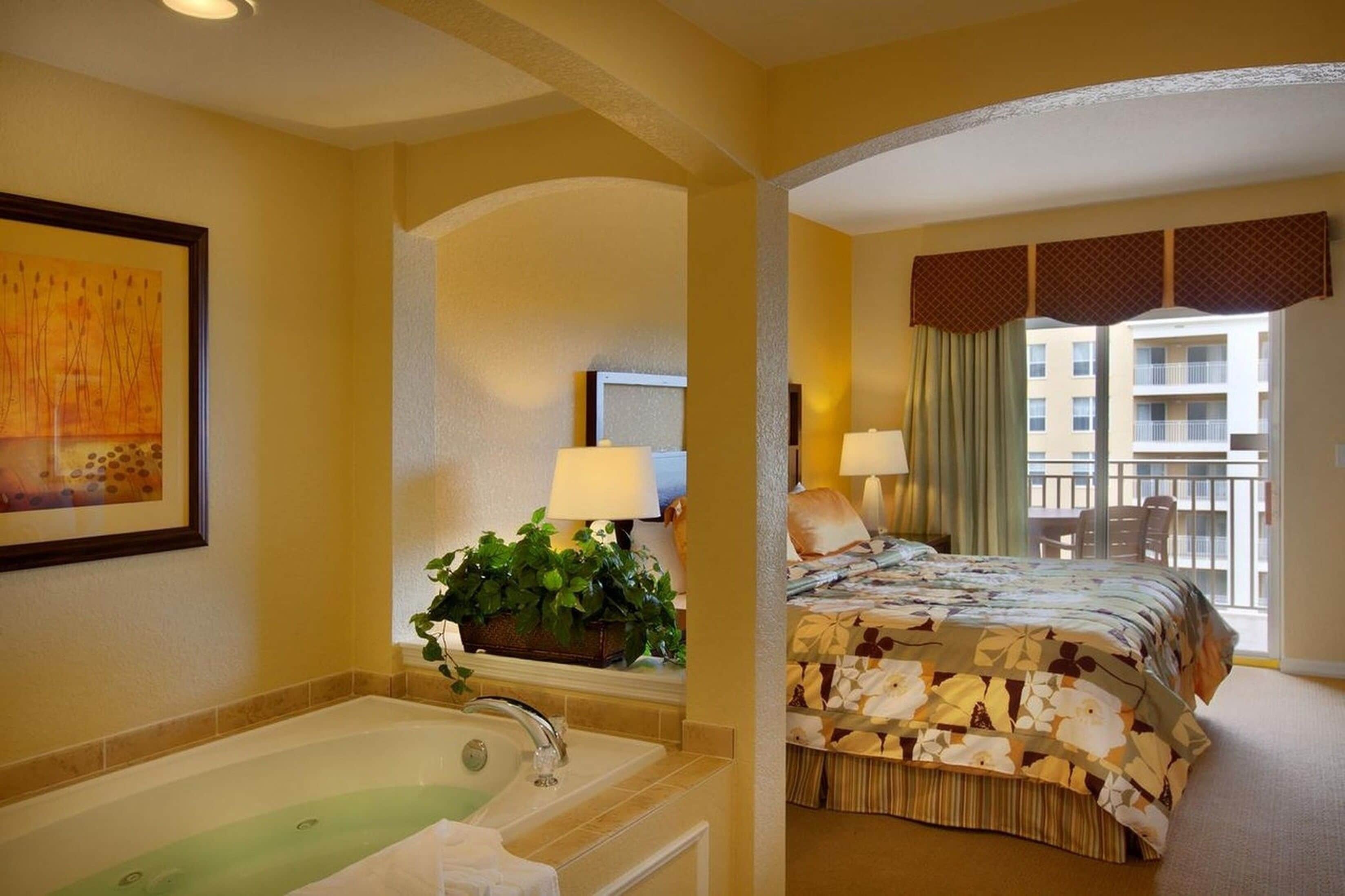 A hotel room with a large bed, a bathroom with a bathtub, a balcony with a view of the building, and a painting on the wall.