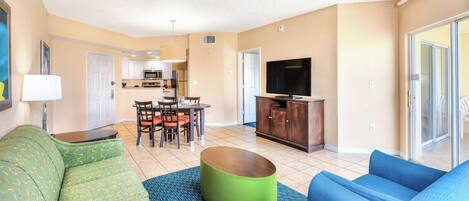 A living room with a sofa, a coffee table, and a television. There is a kitchen area with a dining table and chairs.